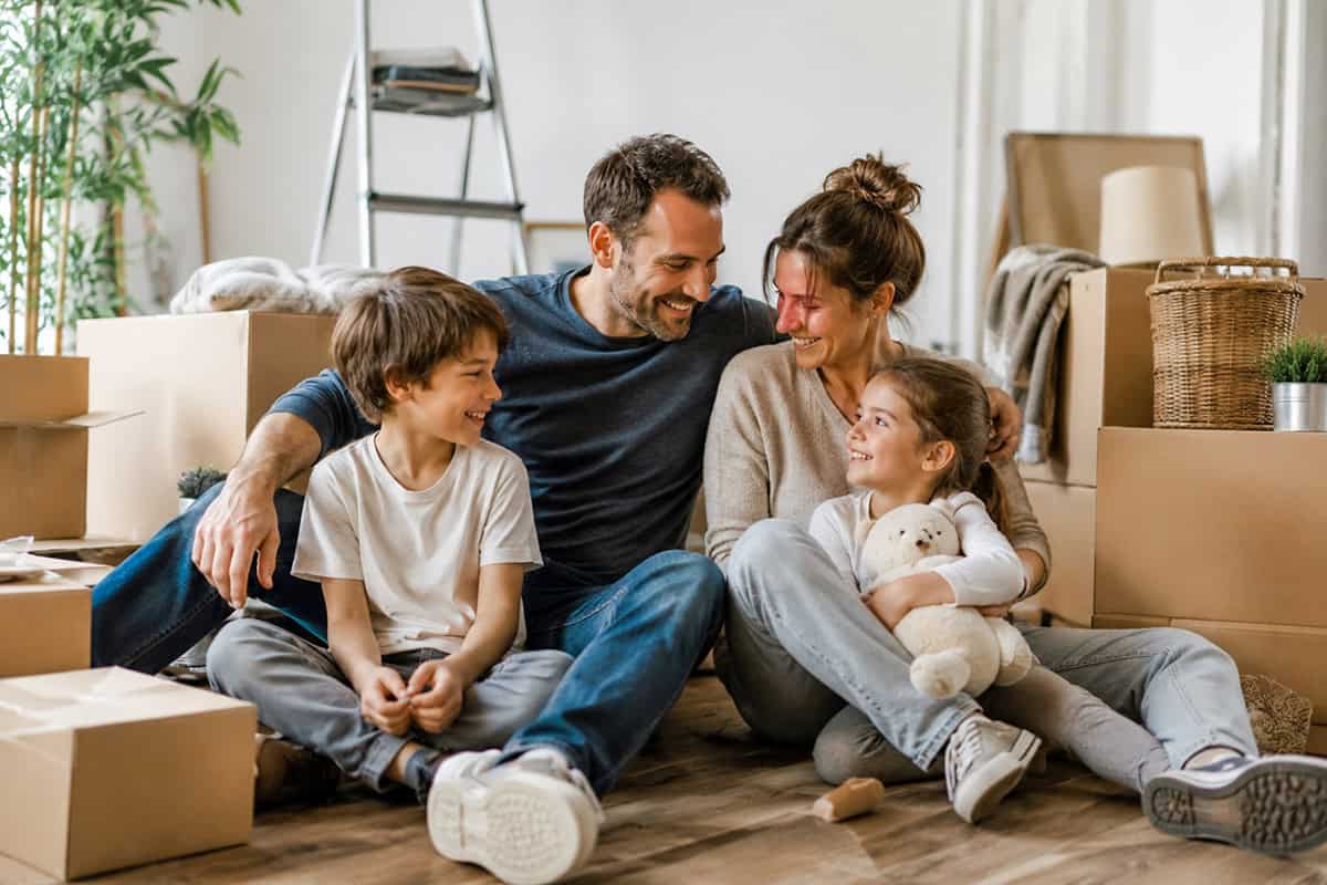 Glückliche Familie mit zwei Kindern sitzt zwischen Umzugskartons in ihrem neuen Zuhause.